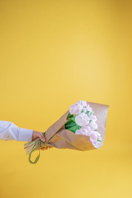 A person in a white shirt holds a bouquet of fresh, light pink tulips wrapped in beige paper with a green ribbon against a vibrant yellow background. The tulips have smooth, slightly glossy petals and
