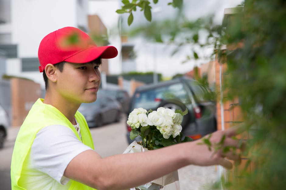 A young delivery person wearing a red cap and a yellow high-visibility vest is holding a bouquet of white hydrangeas, partially wrapped in white paper, with green stems and petals visible. The arrange