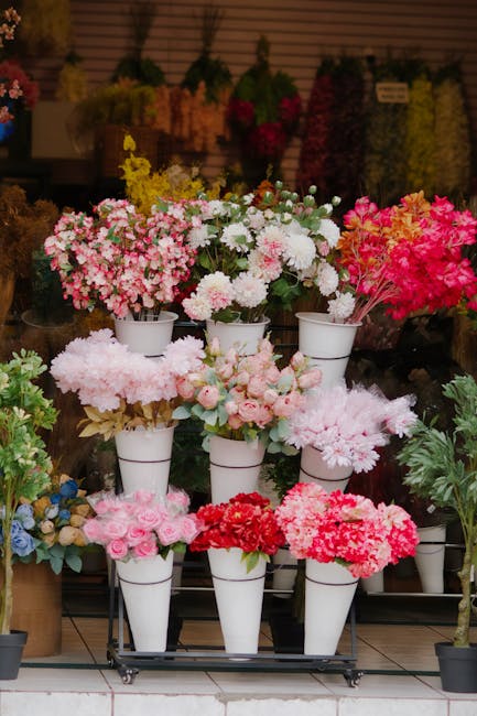 An arrangement of various fresh flowers including pink and white cherry blossoms, roses, and carnations displayed in white ceramic pots on a wheeled stand at a florist shop. The flowers are vibrant, w