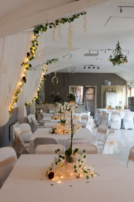 A beautifully decorated indoor venue featuring multiple round tables adorned with white tablecloths and elegant floral centrepieces, including white roses and greenery. Soft ambient lighting is provid