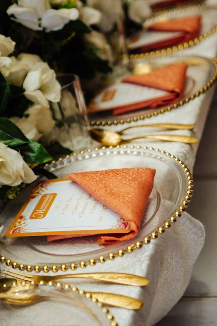 A close-up of an elegant table setting featuring a floral arrangement with white roses and greenery, placed alongside crystal glasses, gold cutlery, and orange napkins folded into triangles on transpa