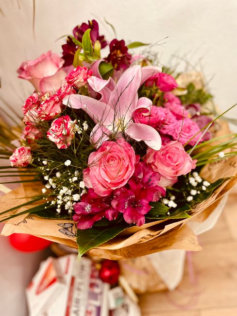 A vibrant floral bouquet featuring a mix of pink roses, pink and white striped carnations, and deep burgundy lilies, arranged with lush green foliage and delicate white baby's breath. The flowers are 