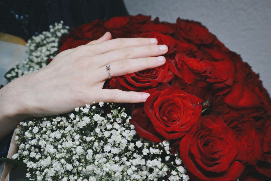 A close-up of a hand with a delicate ring resting on a bouquet of fresh red roses with velvety petals, complemented by white baby's breath flowers with tiny clustered blooms. The bouquet is arranged i