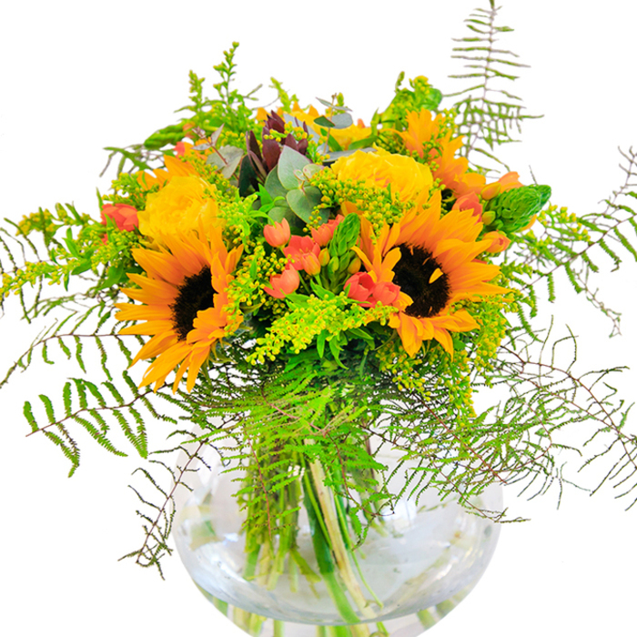 The first impression of this resplendent flower arrangement is one of joyous abundance, crafted with a keen eye for detail tailored to the natural beauty of South Woodford. Dominating the bouquet are glowing sunflowers whose golden petals and bold brown centers evoke the promise of late summer light flooding across Churchfields or the green stretches of Elmhurst Gardens. Nestled beside them, buttery yellow garden roses open in intricate spirals, their soft layers inviting a closer look while lending a sense of refined luxury. Interspersed with these, glossy red Hypericum berries appear as sparkling accents-like the last of summer's berries found in local hedgerows-punctuating the bouquet with vibrancy. The lush composition is softened by trailing fern fronds and delicate, pale green fillers-possibly goldenrod or solidago-each lending subtle movement, their shapes mimicking the gentle swaying of grasses along Tanners Lane. Pale eucalyptus leaves, tinged with blue, peek out from the greenery, their cool tone and faint fragrance adding to the arrangement's depth. The stems, straight and lively, are submerged in crystal-clear water within a rounded glass vase, which provides both a modern touch and a glimpse into the fresh composition held together beneath the blooms. Designed for moments of celebration or everyday appreciation, this bouquet is perfect for bringing the brightness and natural elegance of South Woodford indoors-a testament to the artistry and pride of expert local floristry.
