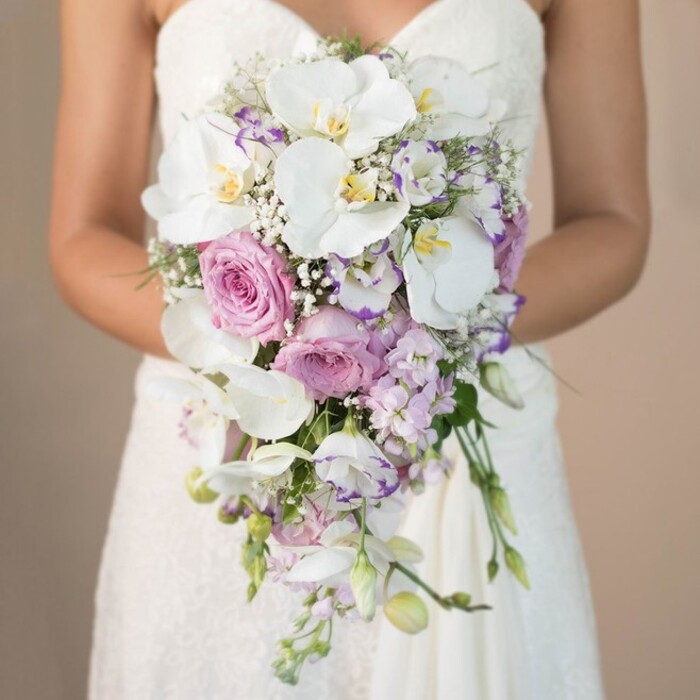 A striking, vertically-oriented image evokes the elegance and romance of a South Woodford wedding, highlighting a bride as she gently holds a luxurious, cascading bouquet. The scene is bathed in soft, natural light-a nod to the tranquil atmosphere found near the River Roding-while a neutral beige background allows the intricate details to stand in full relief. The bouquet itself is a masterpiece of design, shaped in an elongated teardrop that flows like a waterfall. Velvety white phalaenopsis orchids, with distinctive yellow and blush lavender centers, lead the arrangement, their broad petals lending opulence and grace inspired by the floral displays at local events in South Woodford. Nestled alongside, plush garden roses in pastel pink hues, their tightly layered petals echoing classic romance, contrast with smaller blooms of white and pale lavender lisianthus-some petals edged in a richer purple reminiscent of twilight colors over Elmhurst Gardens. Sprigs of tiny white gypsophila float amid the larger blossoms, bringing airy lightness, while discreet hints of fresh green foliage add a subtle, natural volume. The bride's gown, elegantly strapless, features delicate lacework along the bodice and skirt, a timeless style beloved by South Woodford brides. Her gentle touch on the bouquet draws attention to the skilled artistry of local florists and provides inspiration for couples seeking a lavish bridal statement. This image captures the mood of joyful anticipation and refined celebration, offering a vivid vision of wedding florals suitable for same-day delivery or bespoke design in South Woodford.